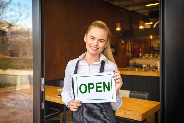 オープン サインの中の人々 を招いて新しい小さな家族経営のレストランの入り口で傾いている若い女性起業家を保持しているレストランのドアに立っている笑顔の所有者の肖像画 - 写真・画像