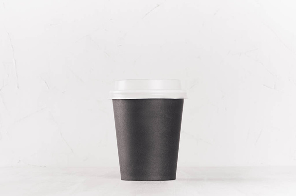 Coffee mockup -  blank black paper cup with white cap on white wood table, coffee shop interior. Modern elegant concept for branding identity, advertising, design. - Photo, Image
