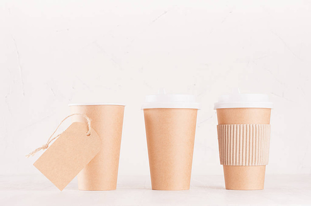 Coffee packing mockup - set of three craft brown paper cups with white caps, cup holder and blank label on white wood board, coffee shop interior. Modern elegant concept for branding identity, advertising, design. - Foto, imagen
