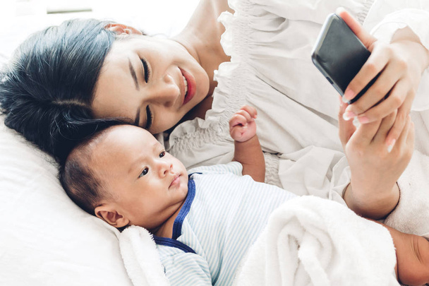 Äiti, jolla on vauva käyttää älypuhelinta valkoisessa makuuhuoneessa.Rakkaus perheen käsite
 - Valokuva, kuva