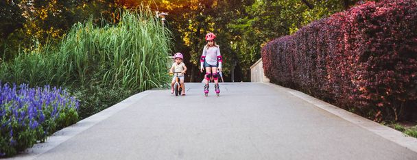 Két kislány lovagol a görgők és runbike nyáron parkban. Gyermek viselése védő párna és biztonsági sisak biztonságos lovagolni. Aktív szabadtéri sport gyerekeknek. Testvérek már versenyben. Nyári sportfelszerelés - Fotó, kép