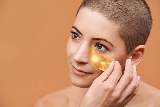 Smiling mid 30s woman applying gold collagen eye mask. Photo of attractive caucasian woman with shaved head and healthy skin isolated on beige background. Beauty products concept. - Fotografie, Obrázek