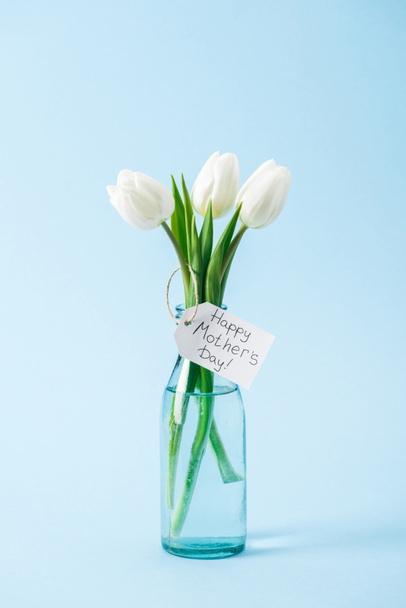bouquet of white tulips in glass vase with happy mothers day greeting label on blue background - Fotoğraf, Görsel