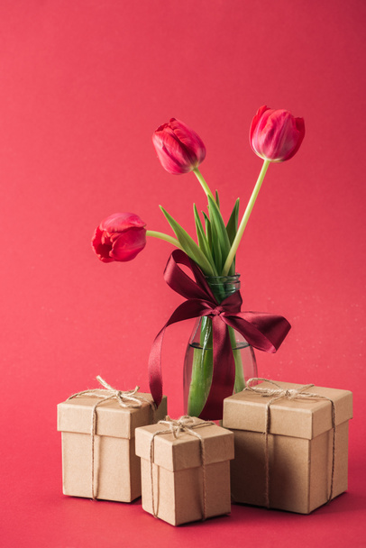 bouquet of red tulips with red satin bowl in glass vase and gift boxes on red background - Fotó, kép