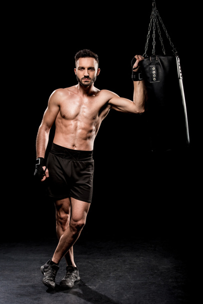 guapo boxeador de pie con las piernas cruzadas cerca del saco de boxeo sobre fondo negro
 - Foto, Imagen