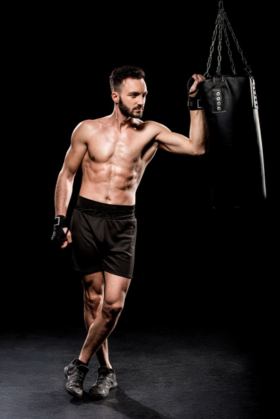 shortless bokser permanent met gekruiste benen in de buurt van bokszak op zwarte achtergrond - Foto, afbeelding