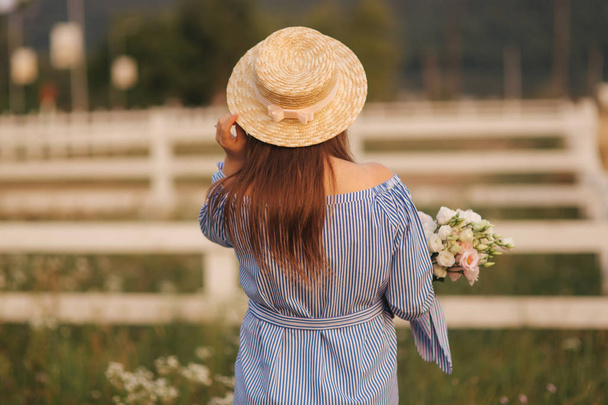 Back view of Beautiful lady. She hold a bouquet of flowers and smile. Background of farm - Foto, immagini