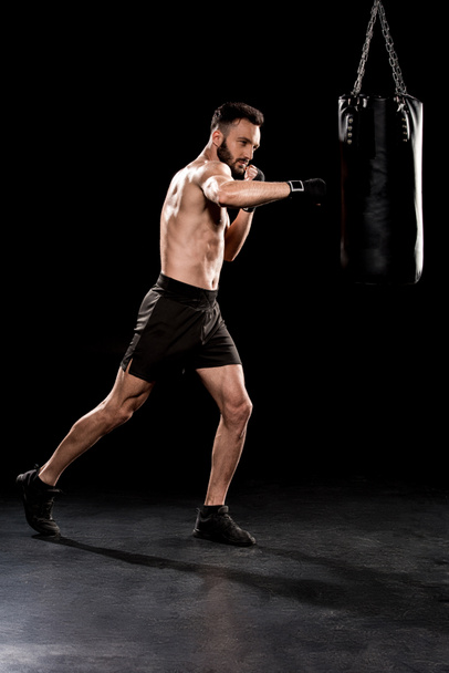 shortless bearded man kicking punching bag  on black background - Foto, Imagem
