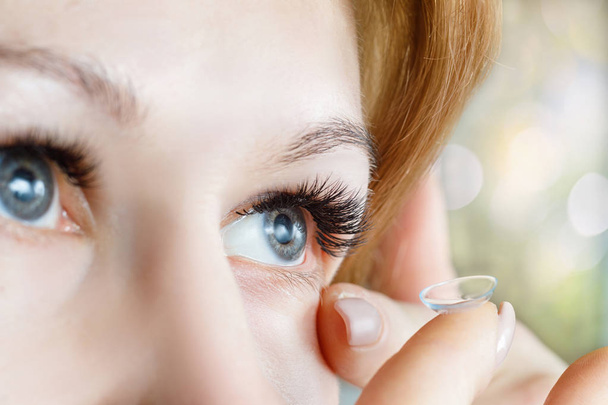 A closeup of a young woman inserting a transparent contact lens into her eye at blurred background. The concept of ophthalmological poor sight correction. - Photo, Image