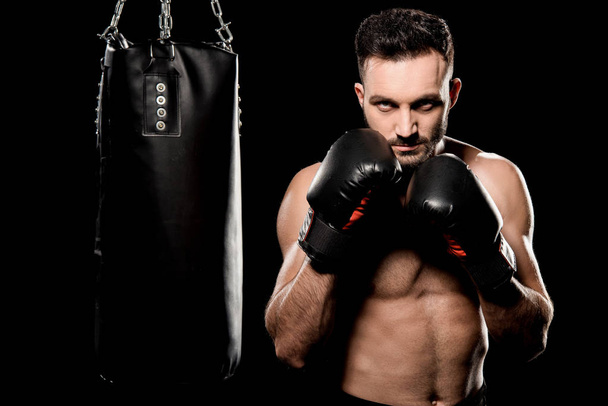 sportovec v Boxerské rukavice v boxerské póze poblíž boxovací pytel izolované na černém pozadí - Fotografie, Obrázek