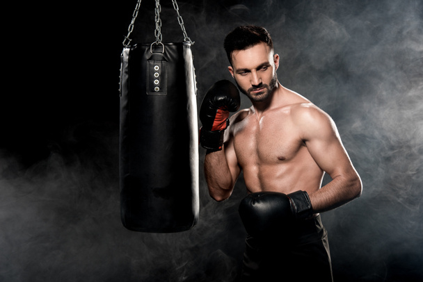 muscular sportsman in boxing gloves standing near punching bag on black with smoke - Φωτογραφία, εικόνα