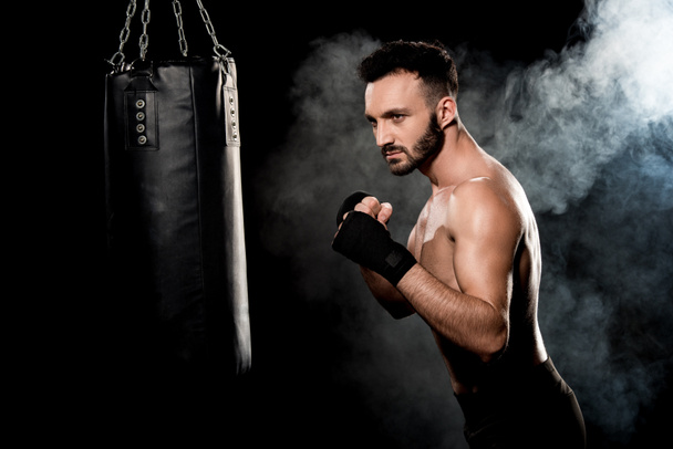 atleta muscular de pie en pose de boxeo y mirando el saco de boxeo en negro con humo
 - Foto, Imagen