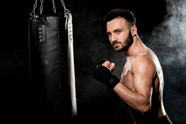 muscular man standing in boxing pose and looking at camera near punching bag on black with smoke - Foto, immagini