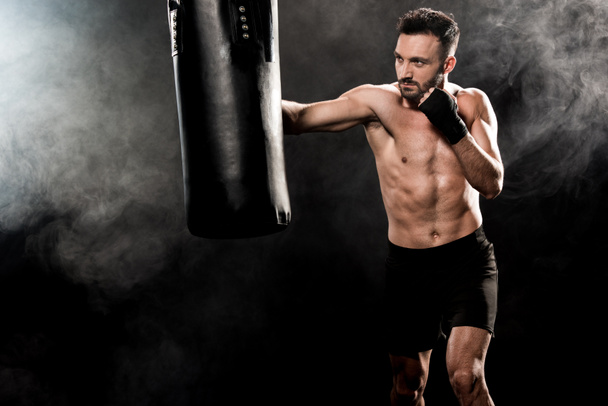 guapo atlético boxeador ejercicio con saco de boxeo en negro con humo
 - Foto, Imagen