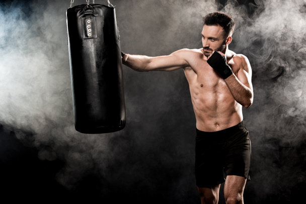 boxeador atlético sin cortocircuito haciendo ejercicio con saco de boxeo en negro con humo
 - Foto, Imagen
