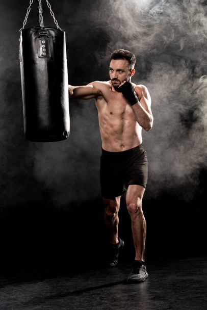 boxeador atlético sin cortocircuito pateando saco de boxeo en negro con humo
 - Foto, Imagen