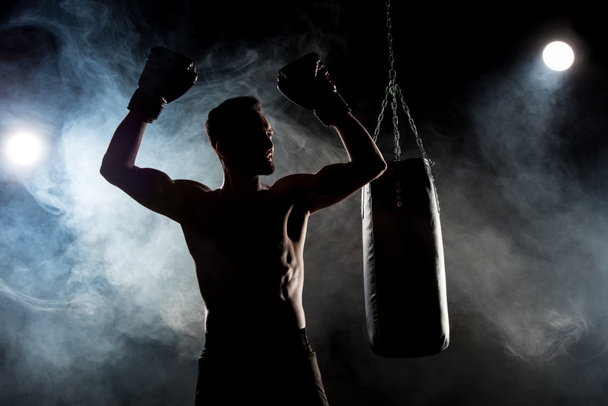 silueta de atleta muscular en guantes de boxeo con las manos por encima de la cabeza en negro con humo
 - Foto, Imagen