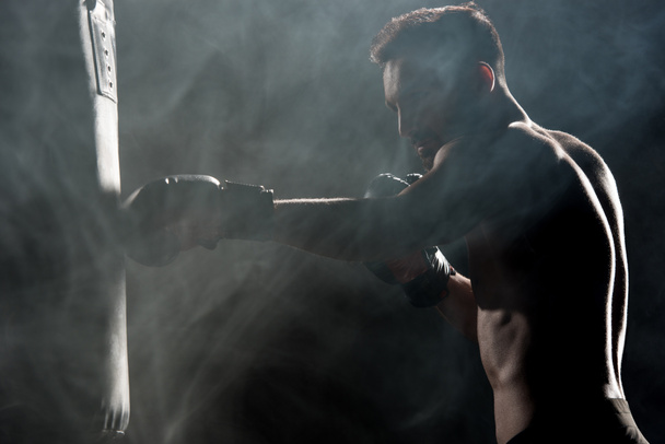 силуэт мускулистого спортсмена в боксёрских перчатках, бьющего боксерскую сумку по черному с дымом
 - Фото, изображение