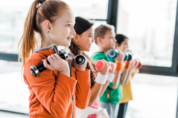 Улыбающиеся дети в спортивной одежде тренируются с гантелями
 - Фото, изображение