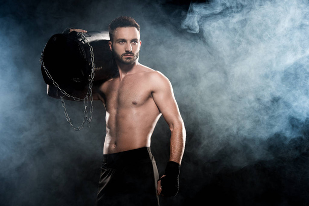 shortless boxer holding punching bag on shoulder on black with smoke - Fotó, kép