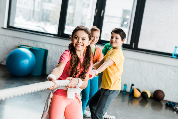 Улыбающиеся дети в спортивной одежде тянут веревку в спортзале
 - Фото, изображение
