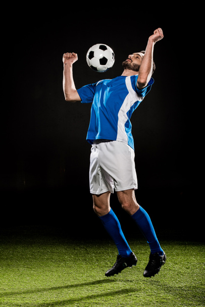 handsome football player in uniform jumping with ball isolated on black - Zdjęcie, obraz