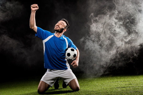 cheerful football player celebrating victory and holding ball on black with smoke - Photo, Image