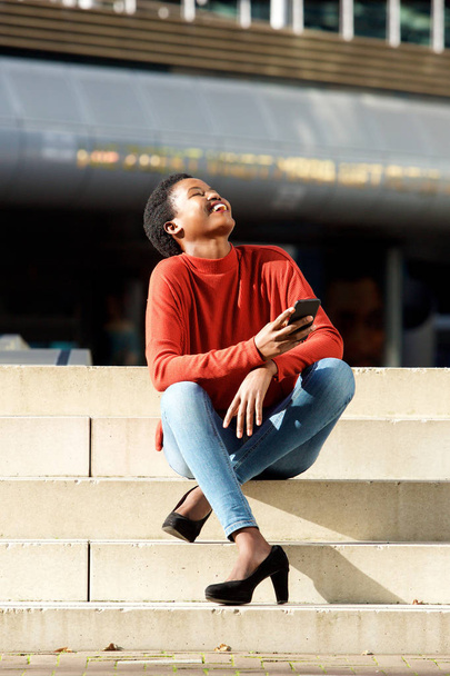 Retrato de cuerpo completo de la joven mujer africana feliz sentada afuera en la ciudad y riendo
 - Foto, Imagen
