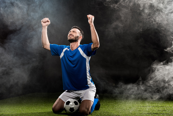 handsome football player celebrating victory while sitting on grass on black with smoke - Photo, Image