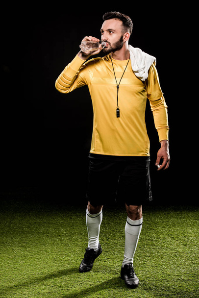 bearded referee drinking water from bottle and standing with towel on shoulder isolated on black - Foto, Imagem