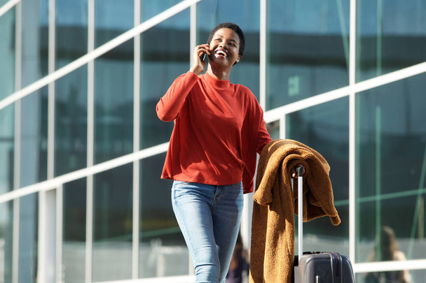 Portret van mooie jonge zwarte vrouw lopen in de stad met mobiel en koffer in de hand - Foto, afbeelding