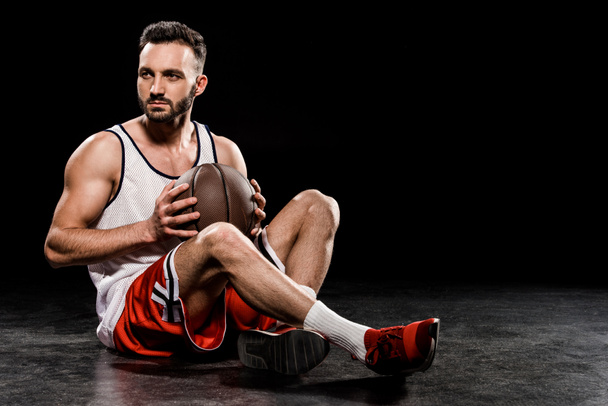 muscular basketball player sitting with ball on black background - Фото, изображение