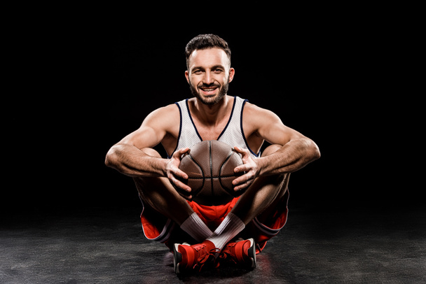 fröhlicher Basketballspieler sitzt mit Ball auf schwarzem Hintergrund - Foto, Bild