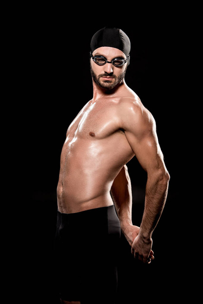 muscular swimmer posing in swimming cap and goggles isolated on black  - Photo, Image