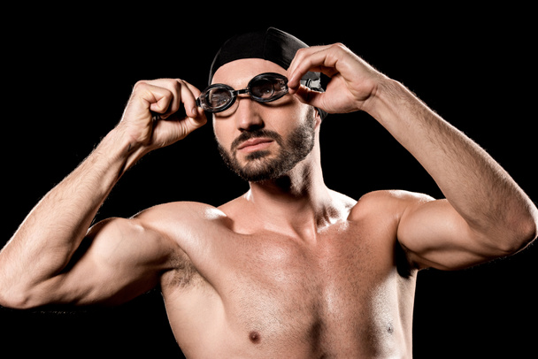 athletic sportsman standing in swimming cap and touching goggles isolated on black  - Photo, Image