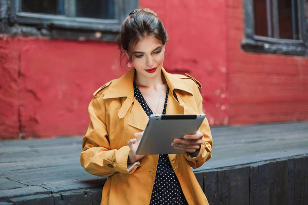 Young attractive woman in orange trench coat and black polka dot dress thoughtfully using tablet spending time in old courtyard - Фото, изображение