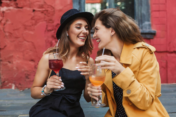 Young beautiful laughing girls holding cocktails in hands happily looking at each other spending time together in old cozy courtyard of cafe - Photo, Image