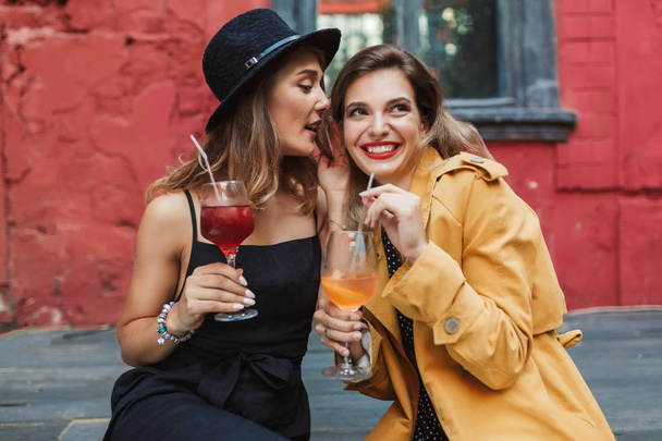 Young attractive woman in black dress and hat thoughtfully talking gossips to pretty smiling lady holding cocktails in hands spending time together in old courtyard of cafe - Photo, Image