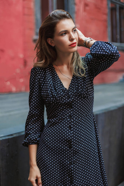Young attractive pensive woman in black polka dot dress with red lips thoughtfully looking aside spending time in old courtyard - Foto, Imagen