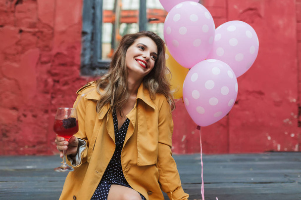 Giovane donna attraente sorridente in trench arancione che tiene in mano cocktail in sogno guardando da parte con palloncini rosa vicino nel vecchio cortile del caffè
 - Foto, immagini