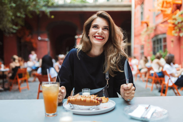 Vrolijke jongedame in zwarte jurk bedrijf vork en mes in handen met voedsel op tafel kijken vreugdevol tijd opzij doorbrengen in gezellige binnenplaats van café  - Foto, afbeelding