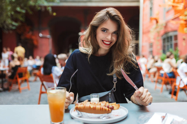 Jonge mooie lachende vrouw in zwarte jurk bedrijf vork en mes in handen met voedsel op tafel kijken gelukkig tijd in de camera doorbrengen in gezellige binnenplaats van café  - Foto, afbeelding