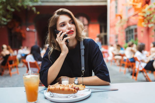 Mladá krásná žena v černých šatech sedí u stolu s jídlem na mobil zasněně dívá stranou v útulné nádvoří kavárny  - Fotografie, Obrázek