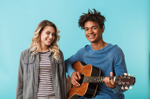 Happy young multiethninc couple together, playing a guitar isolated over blue background - Fotografie, Obrázek