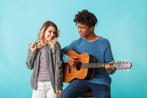 Happy young multiethninc couple together, playing a guitar isolated over blue background - Foto, Imagen
