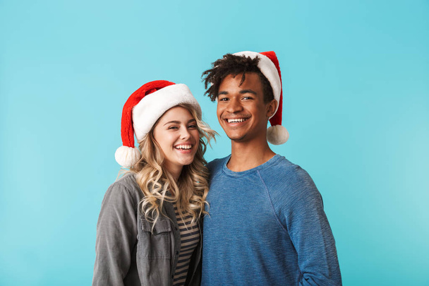 Happy multiracial young couple wearing christmas red hat isolated over blue background - Foto, Imagem