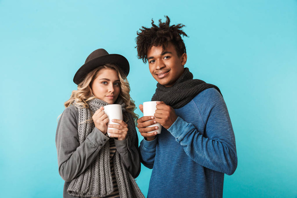 Multiracial teenage couple wearing winter clothes standing isolated over blue background, holding cups of tea - Zdjęcie, obraz