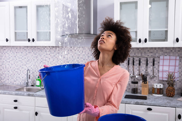 Close-up Of A Worried Young Woman Collecting Water Leaking From Ceiling In The Blue Bucket - Фото, зображення