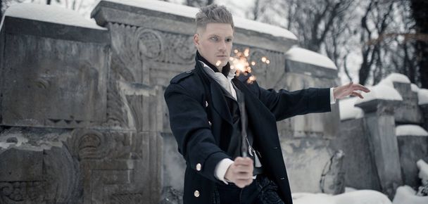 Young man in image of black magician makes a spell using magic wand next to crypt. - Photo, Image