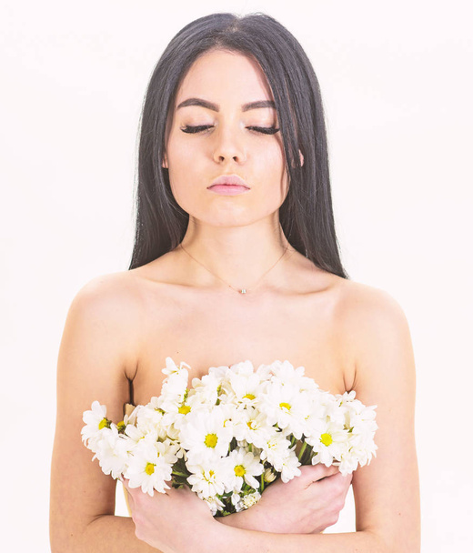 Skin care concept. Girl on calm face stands naked and holds chamomile flowers in front of chest. Woman with smooth healthy skin looks attractive. Lady covers breasts with flowers, isolated on white. - Foto, afbeelding